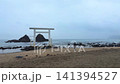 A couple poses under a torii gate on Itoshima's Sakurai Futamigaura beach as an assistant holds an umbrella. Waves crash against the sacred couple stones and shimenawa rope. Cinematic Fukuoka coast. 141394527