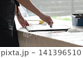 Woman creating textured artwork on canvas at home spreads white plaster paste with a palette knife at work table near a large window. Concept of DIY wall decor, handmade art and creative hobby. 141396510
