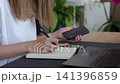 Woman planning home budget with phone, writing notes in a notebook with euro cash and laptop on the table. Concept of household expenses and bills. 141396859