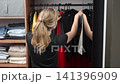Woman choosing outfit in a wardrobe holds black top and red skirt by the mirror, with hanging clothes behind her. Concept of getting ready and outfit choice. 141396909