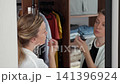 Woman applying mascara in front of a mirror, with clothes and shelves in the background. Concept of eye makeup, beauty and getting ready 141396924
