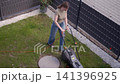 Woman mows grass with an electric lawn mower in a small fenced garden. Concept of lawn care, gardening and yard maintenance. 141396925