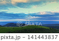 Wind turbines stand on a hilltop as clouds gather in the sky and the sun sets in the distance over a green landscape 141443837