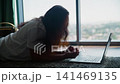 Woman Lying on Floor Viewing Laptop by High Rise Window 141469135