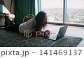 Woman lying on floor working on laptop in high rise apartment with city view 141469137