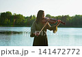 Woman playing violin by a tranquil lakeside surrounded by lush green forest, enjoying peaceful 141472772