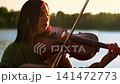 Woman playing violin by serene lakeside at sunset, focusing on emotional musical performance 141472773