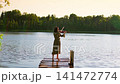 Young woman playing violin on wooden pier over calm lake at sunset, surrounded by peaceful forest 141472774