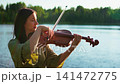 Woman playing violin by tranquil lakeside in soft natural evening light, creating peaceful outdoor 141472775