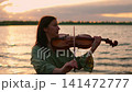 Violinist performing by tranquil lakeshore at golden sunset, serene woman playing classical music 141472777