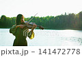 Young woman playing violin by tranquil lakeside at sunset, silhouetted against shimmering water and 141472778