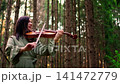 Woman playing violin in tranquil forest, creating peaceful music among tall trees with soft natural 141472779