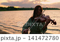 Woman playing violin by tranquil lakeside at sunset, silhouetted against glowing sky and gentle 141472780
