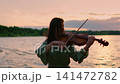 Woman playing violin by tranquil lakeside at colorful sunset, creating serene music as soft evening 141472782