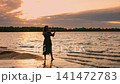 Woman playing violin at tranquil lakeside shore during golden sunset, standing barefoot in gentle 141472783