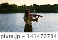 Young woman playing violin by tranquil lakeside at golden sunset, creating peaceful music in nature 141472784