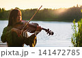 Young woman playing violin by tranquil lakeside at golden sunset, creating serene music in nature 141472785