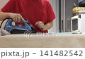 Hands use a steam iron to smooth a checked towel on an ironing board in a home kitchen. 141482542