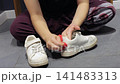 A person wipes the sole edge of a white sneaker with a red sponge on a gray tile floor. 141483313