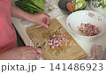 In the kitchen, a woman is busy preparing a salad by chopping cabbage, cucumber, and radish. She adds fresh herbs to create a tasty dish for lunch or dinner 141486923