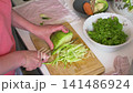 In a kitchen, a woman is chopping cabbage on a cutting board. She prepares ingredients for a healthy salad using fresh vegetables from her countertop 141486924