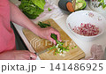 A woman is chopping onions on a wooden cutting board in her kitchen. Fresh vegetables are on the counter, and she is preparing for a salad 141486925