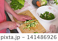 In a kitchen, a woman is busy slicing cucumbers on a cutting board. She prepares fresh ingredients for a salad. Other vegetables are visible in bowls nearby 141486926