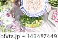 A woman prepares a cold summer salad called okroshka. She combines cooked potatoes, radishes, cucumbers, chopped chicken, fresh greens, eggs, and kefir in a bowl 141487449