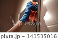 Close-up cropped shot of person wearing blue rubber glove cleaning bathroom mirror with orange cloth. Housewife, maid doing housework, wiping glass surface. Household chores and hygiene concept. 141535016