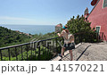 Cervo, liguria, italy. June 22, 2025. Female tourist with a backpack admiring the stunning panoramic view of the mediterranean sea from a balcony in the historic village of cervo 141570221