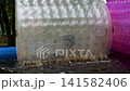 Transparent rolling tube in motion. Water splashes around the tube. Pink tube nearby. Outdoor grassy area background. No animals or plants present 141582406