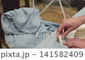 Hands iron fabric on a table. Fabric is light blue and wrinkled. Iron glides smoothly over surface. Hands are steady and precise. Background shows draped cloth. Scene is calm and focused 141582409