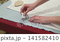 Hands mark fabric with a white chalk stick. Fabric rests on a red ironing board. Paper pattern lies beneath the fabric. Hands show precision in marking fabric edges. Work surface holds a smooth stone 141582410