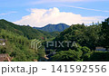 A summer scene of a rural mountain village with towering cumulonimbus clouds; a natural landscape of blue sky and lush greenery. 141592556
