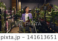 Woman choosing plants from shelves with cart partially filled. Relaxed shopping mood, browsing the store 141613651