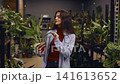Young woman holding potted plant smiling inside urban plant shop. Browsing shelves of tropical houseplants and terracotta pots, warm ambient 141613652