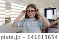 Happy young girl explores eyewear options inside modern shop. Bright smiling girl reviews and fits glasses in contemporary optical boutique 141613654