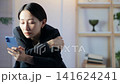 A middle-aged woman with a gloomy expression looks at her smartphone. Worried, depressed, disappointed, melancholic, looking at social media and videos, lonely, stressed. 141624241