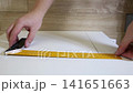 Working hands use a yellow metal ruler and a marking gauge to score a straight line on a white laminate board. Precision measurement for woodworking. 141651663