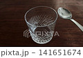 Metal teaspoon taps on empty crystal drinking cup. Clear glass with textured pattern on dark brown wooden table surface, ready for drink preparation or serving beverage 141651674