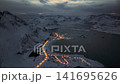 Aerial night view of snowy coastal village on a fjord with winding illuminated road 141695626