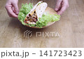 Person holding a wooden plate with a meat shawarma wrap and fresh green lettuce. The savory wrap is filled with grilled meat and red sauce, served on a rustic wooden tabletop background. 141723423