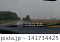 Rainy rural road seen through windshield with wipers, approaching car, fields, trees, and gray sky 141734425