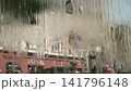 A dense, continuous stream of water flows from the edge of a fountain, shot from inside, with an old architectural building in the background, in the city center, on a hot summer day. 141796148