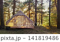 Glamping tent glowing at sunset among tall pine trees inside quiet forest campsite, warm interior light, soft sky behind branches, rustic outdoor retreat in nature 141819486
