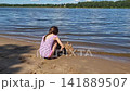 A little girl is building a sandcastle on a hot summer day by the riverbank. 141889507