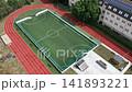 Aerial drone shot of a modern school sports ground in Hungary featuring a fenced artificial turf football pitch and red running tracks. 141893221