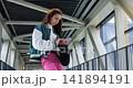 Young woman checking smartphone on walkway, varsity jacket and pink pants, standing by metal railing in glass corridor, scrolling messages with focused expression, urban commuter mood and soft 141894191
