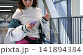 Young person in varsity jacket enjoying phone and candy outside bridge. Youthful individual dressed in varsity clothing engaging with mobile device beside bridge while eating lollipop 141894384