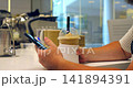 Hand holding cold drink and smartphone at cafe table, with iced beverage in takeaway cup, black straw, and person checking phone inside modern coffee shop interior with clean design. 141894391